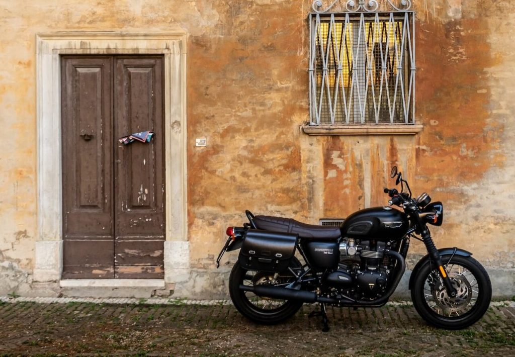 Un'immagine di una moto café racer nera posizionata contro un muro giallo, con una porta di legno e una finestra sullo sfondo.